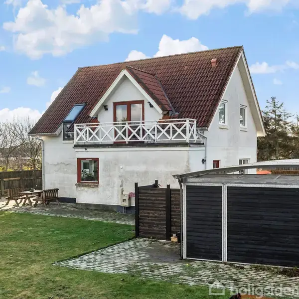 Hus står stille i en have med græsplæne. Bygningen har rødt tag, hvid facade og en balkon. Der er et terrassebord ved siden af en garage.