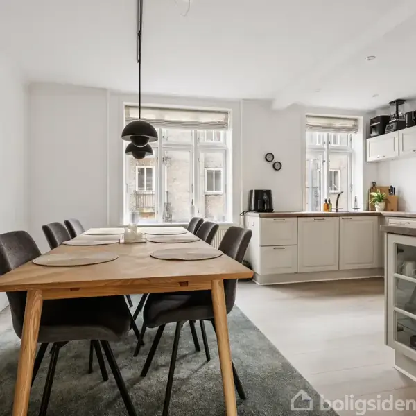 Et spisebord med seks stole står i en lys spisestue. Der er dækket med dækkeservietter. Et sort, moderne loftslys hænger over bordet. I baggrunden ses køkkenelementer.