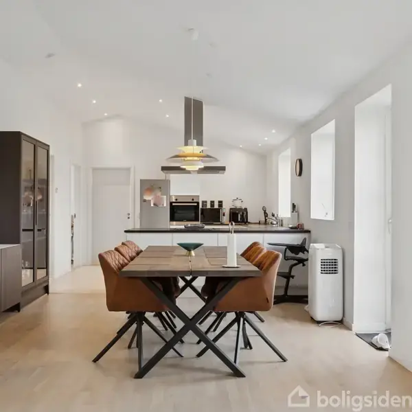 Et spisebord med fire stole står centralt placeret. Baggrunden viser et moderne, åbent køkken med hvide vægge og lofter, indbyggede skabe, og en køkkenø med integreret komfur og emhætte.