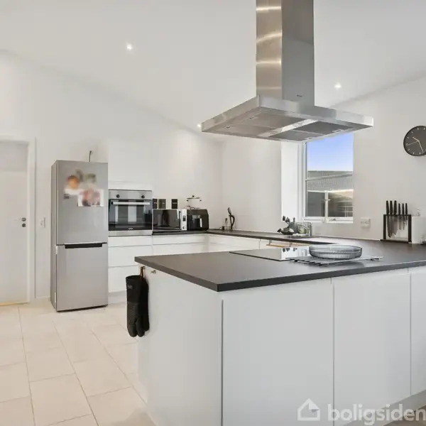 Et moderne køkken med en stor ø i midten udstyret med en emhætte. Hvide skabe dominerer rummet. Et ur hænger på væggen, og køkkenapparater er placeret på bordene.