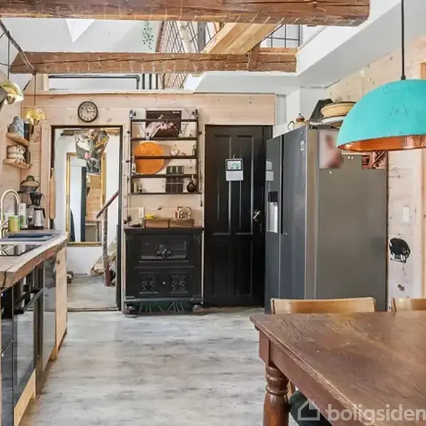 Køkken med træmøbler, hvor der hænger to turkise lamper over et spisebord. Et sort komfur, grå køleskab og en blomsterbukets vase står på bordet.