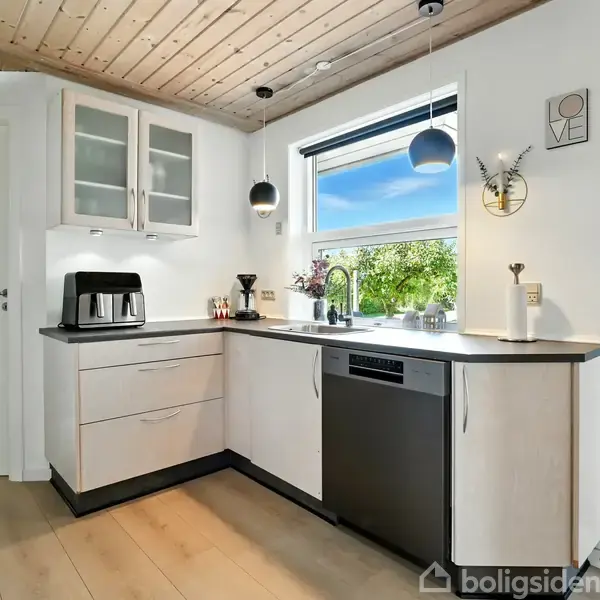 Køkken med hvidt træloft og lyse trægulve. Kaffemaskine står på køkkenbord ved siden af et vindue med udsigt til en grøn have. Dør med teksten "MYNTE".