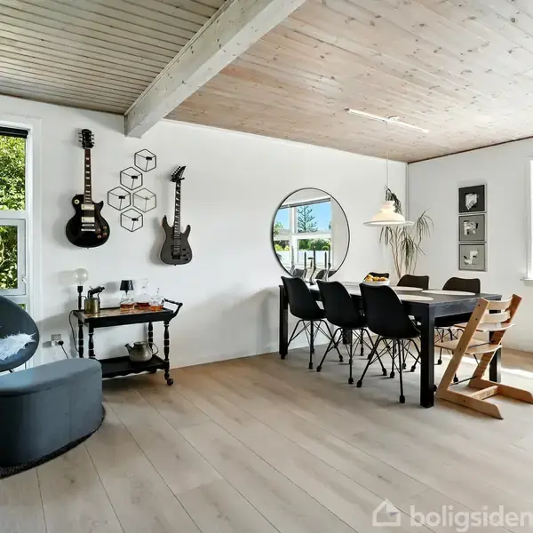 En stue med lyse trægulve har en rund stol, to guitarer på væggen, et rundt spejl, en spisebord med seks stole, og store vinduer, der lukker naturligt lys ind.