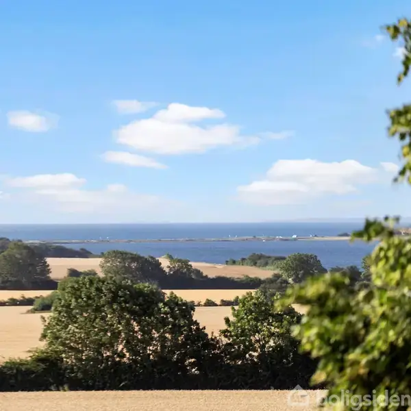 Landskab med udsigt over marker og træer mod en blå himmel og havet i baggrunden. Skyer svæver let på himlen, og der er frodige grønne områder. teksten "boligsiden" er nederst.