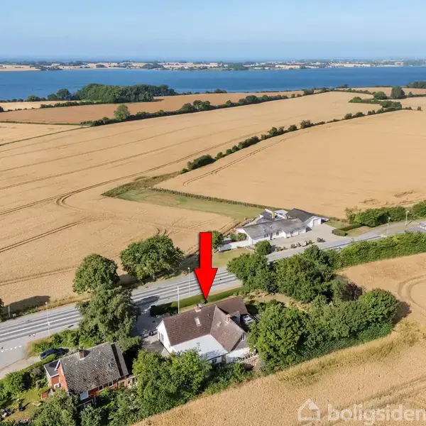 Et hus beliggende blandt åbne marker med en rød pil, der peger på det. Baggrunden inkluderer en vej og en smuk kystlinje med vand, omgivet af grønne områder.