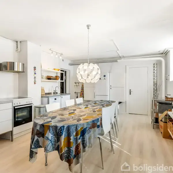 En spisestue med et bord dækket af en farverig dug, omgivet af stole. I baggrunden ses et moderne køkken med ovn og køleskab. En lampe hænger over bordet.