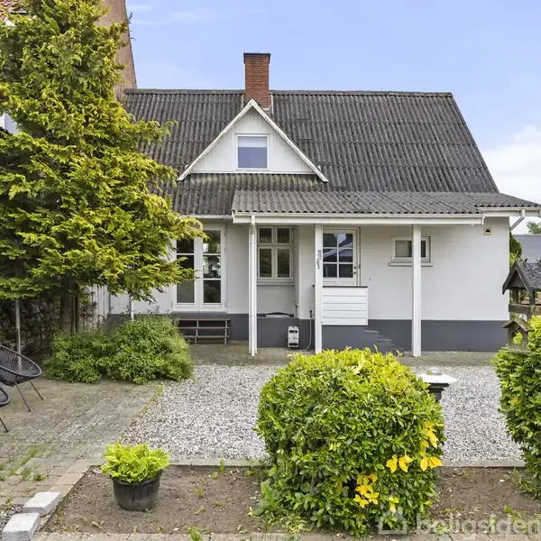 Et hvidt hus med mørkt tag står i en have, omgivet af buske og fliser. Foran huset er der grus og en dør med vinduer på hver side.