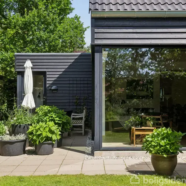 Moderne hus med store vinduer vender mod en terrasse. Terrasse omgivet af grønne planter i potter. Stol og parasol på terrassen. Grønne træer og buske omkranser området.
