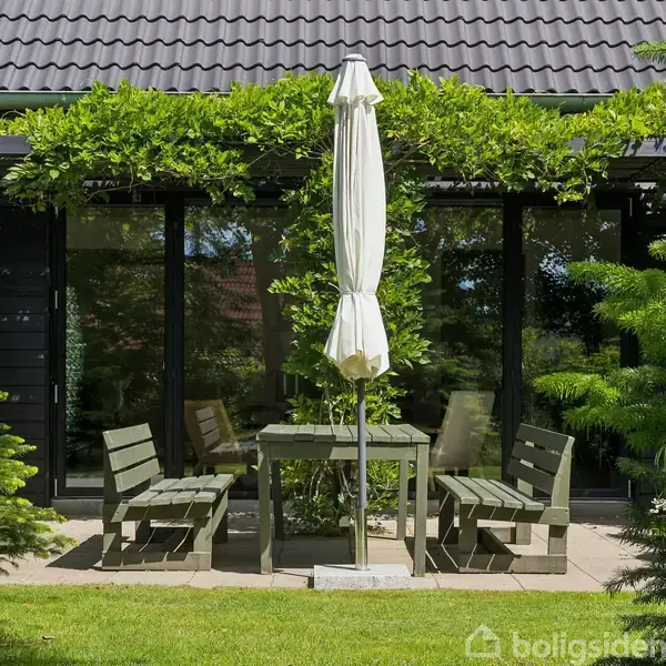 Et sæt udendørs bord og stole står på en terrasse med en uåbnet parasol i midten. Omgivet af grønne træer og buske foran et hus med glasdøre.
