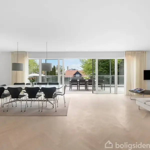 En moderne stue med spisebord og seks stole i midten, vendt mod en stor glasdør, der åbner mod en terrasse med udsigt til træer. Et sofahjørne til højre.