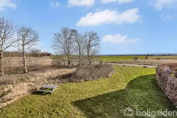 En picnicbænk står på en grøn græsplæne. Baggrunden viser åbne marker og træer under en blå himmel med mange skyer.