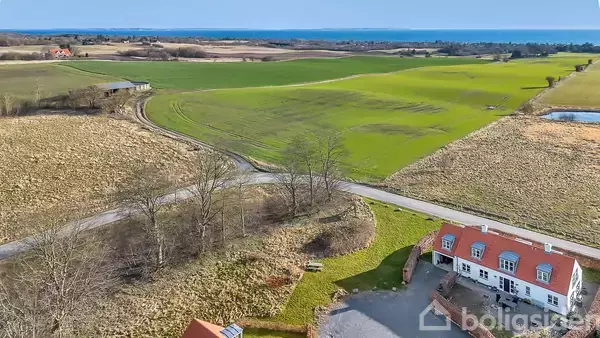 Et hvidt hus med rødt tag står ved en vej i en landlig scene; grønne marker strækker sig ud til horisonten med havet i baggrunden.