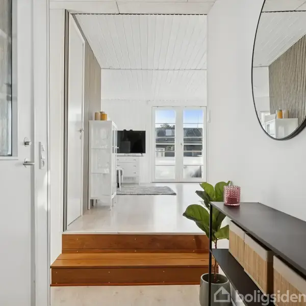 Rundt spejl hænger på en hvid væg over et mørkt sidebord med planter og lys, med trin der fører til en stue. Store vinduer og et tv tydeligt i baggrunden.