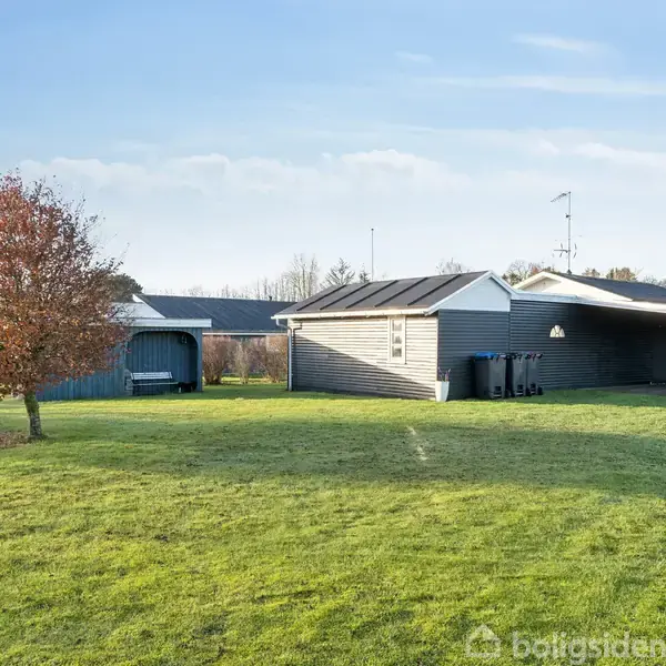 En grå bygning med carport står på en grøn græsplæne. Et ensomt træ ses til venstre, og skovbeklædte bakker udgør baggrunden.