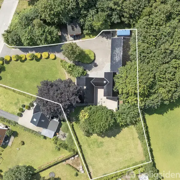 Hus set ovenfra omgivet af træer og grønt græsareal. En lang indkørsel fører til hovedbygningen. Mindre bygninger og en have er synlige i nærheden.