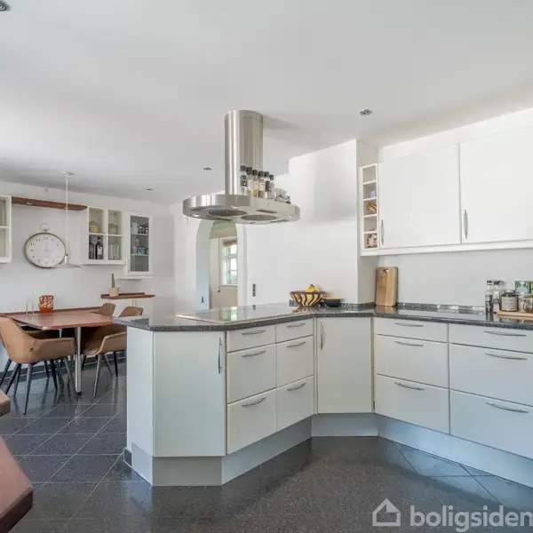 Et moderne køkken med hvide skabe og en central emhætte står i et lyst rum. En træbordplade rammer stålvasken, og spisestuen med bord og stole ses i baggrunden.