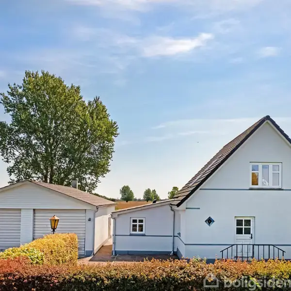 Hvidt hus med mørkt tag står i en landlig have omgivet af græsplæne. Til venstre er en garage med to porte, og store træer står i baggrunden under en blå himmel.