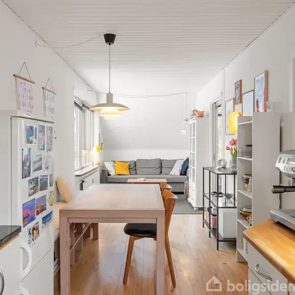 Et køkken-alrum med en kaffemaskine på bordpladen, et træbord med stole, og en grå sofa i baggrunden ved vinduet. Væggene har dekorationer og billeder.