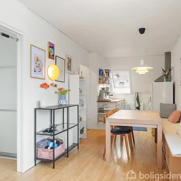 Et køkken med et spisebord midt i rummet, omgivet af stole og en bænk. Til venstre ses en skænk og reol. Væggene har kunst og hylder.