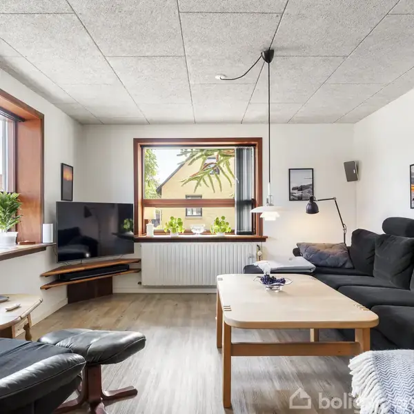 Sort sofa står i stue, komplet med sorte stole omkring et træbord. Et stort vindue lader lys ind, og planter er placeret på vindueskarmen.