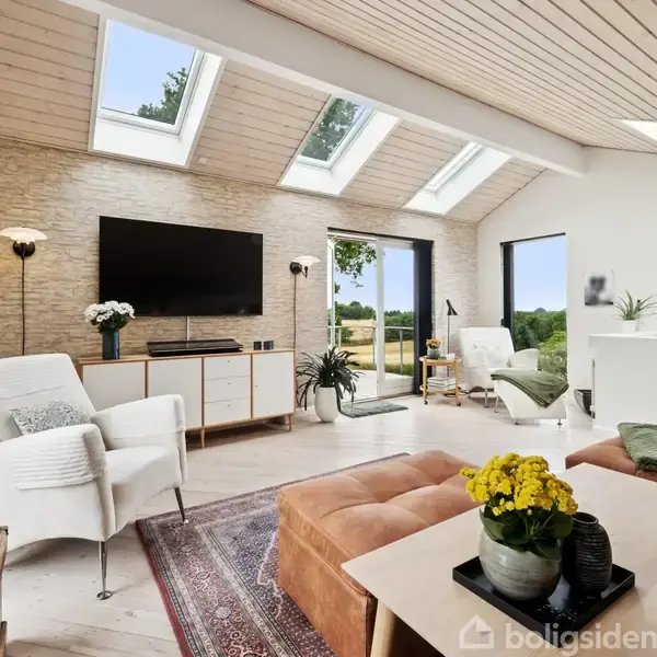 Stue med lyse møbler og sofa, fladskærms-tv på stenmur, store ovenlysvinduer. Lysegul blomst på bord, grøn udsigt udenfor døren. Tæppe dækker trægulv, hyggelig atmosfære.