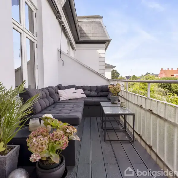 En balkon med en L-formet sofa, små borde og planter i krukker. Balkonen har udsigt til et grønt område med huse i baggrunden under en blå himmel.