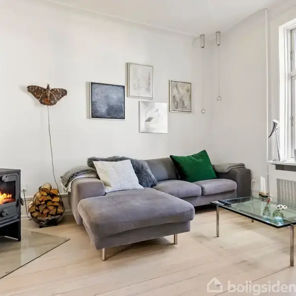 En grå sofa med puder står ved væggen dekoreret med billeder. En brændeovn med ild ved siden af brændestabel. Et glasbord foran, med et vindue til højre.