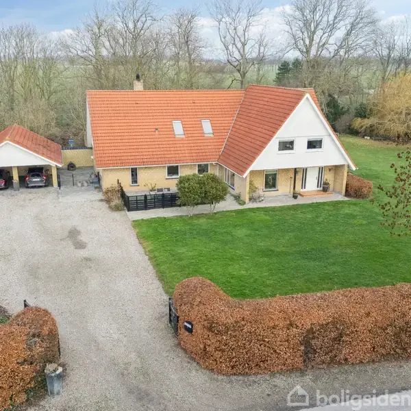 Et gult murstenshus med rødt tag står stille i en frodig have med grønne græsplæner og hække. Til venstre ses en grusindkørsel og en dobbelt carport.