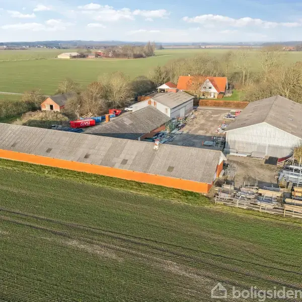 En stor gård med flere længer og bygninger ligger spredt ud i et frodigt, fladt landskab. Omgivet af marker og skove ses gården fra en luftfoto-vinkel.