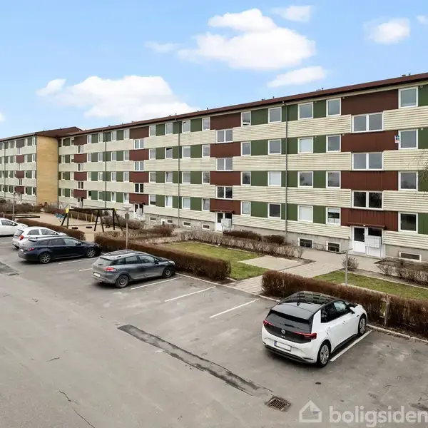 En lang, fleretagers boligblok står stille i et p-hus. Parkeringen er fyldt med biler, og omgivelserne er en klar blå himmel med få skyer.