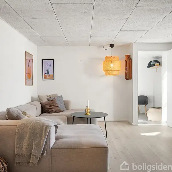 Stue med beige hjørnesofa, pyntepuder, og sofabord med lysestager. Vægge har kunstplakater. En orange lampe hænger fra loftet. Lyse trægulve og en dør fører til et andet rum.