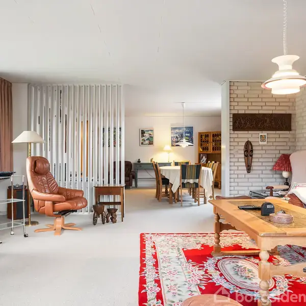 Stue med lænestol og sofabord, hvor en rød orientalsk tæppe pryder gulvet. I baggrunden ses en spiseplads med borde og stole. Stemningsfuld belysning fra en loftslampe.