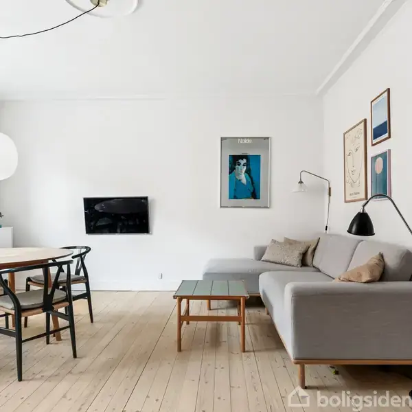 En moderne stue med en grå sofa ved siden af et sofabord. På væggen hænger flere kunstværker og en fladskærm. Rundt spisebord med stole i træ.