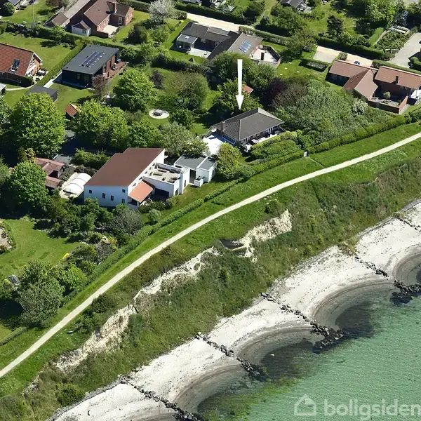 Huse lokaliseret langs en kyst med klipper og strand, omgivet af frodige træer og haver, set fra luften. En sti adskiller husene fra klitområdet ved vandet.