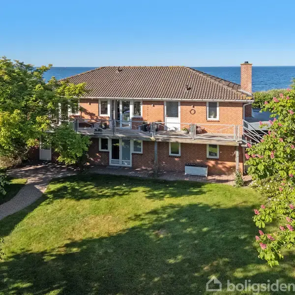 En bro strækker sig over vandet, set fra en kyst med grønne buske i forgrunden, under en klar blå himmel.