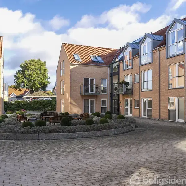 En gårdhave med bord og stole omgivet af lave buske. Bagved står to-tre-etagers murstensbygninger med balkoner og store vinduer. Sollys belyser området, og en stor træ ses i baggrunden.