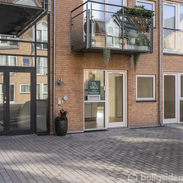 En moderne murstensbygning med altaner og glasdøre. Der er en plakat med teksten "TIL SALG danbolig" på en af glasdørene. Omgivet af en flisebelagt gårdsplads.