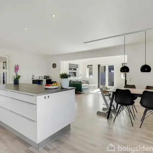 Køkkenø med frugtskål og orkidé står i et åbent, lyst rum. Ved siden af er et spisebord med fire sorte stole, og i baggrunden en stue med sofa.