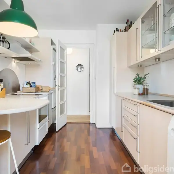 Et lyst køkken med trægulv har en rund spisebar med en grøn lampe. Køkkenet har skabe, en vask, en ovn, og hylder med planter og krydderier.