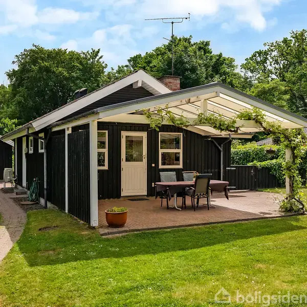 Sort træhus med hvid indgangsdør, omgivet af en græsplæne. En overdækket terrasse foran, møbleret med bord og stole, ligger i en frodig have med flere træer i baggrunden.