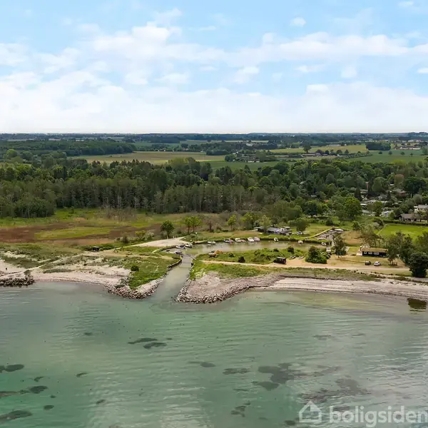 Kystlandskab med en mole, der strækker sig ud i havet. Omgivet af træer og marker, ses en mindre landsby i baggrunden under en blå himmel med skyer.
