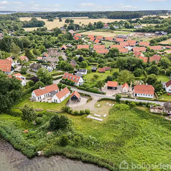 Kystby med røde tage og grønne træer langs en flod. Huse ligger tæt på hinanden med marker i baggrunden. En vej snor sig gennem landskabet.