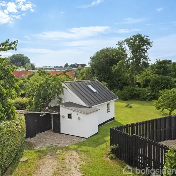 Et lille, hvidt hus med skråt tag står stille i en frodig have omgivet af træer og buske. En grussti fører til huset, og et sort stakit omgiver en del af grunden.