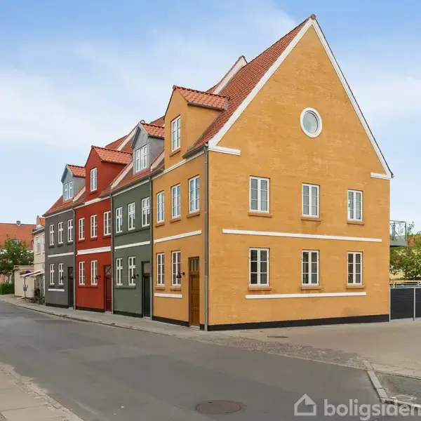 Rækkehuse med farvede facader står stille på en hjørnegade i et roligt boligområde. Husene har røde tegltage og klassiske vinduer under en lys blå himmel.