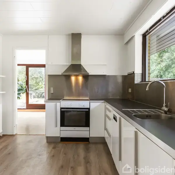 Et moderne køkken med en hvid ovn og emhætte, hvor kogeplader er i brug. Omgivet af hvide skabe og en stor vinduesrude, der lukker grønne omgivelser ind.