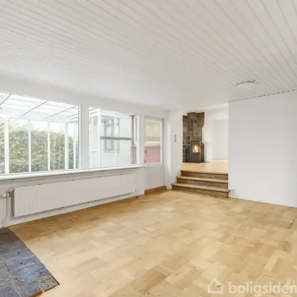 Stue med trægulv og hvidmalet loft, store vinduer til venstre giver naturligt lys. En pejs på murstensvæg står forenden af rummet, tæt på en trappe.