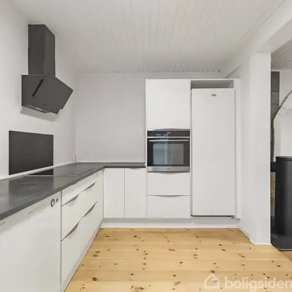 Et moderne køkken med hvide skabe og skuffer, sort emhætte, og køleskab ved siden af en indbygget ovn. Trægulv og hvid loft i en lys rum.