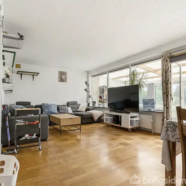 En stue med en hjørnesofa står i et lyst rum, mod en væg med et kunstbillede. Et stort TV står på et lavt bord ved vinduet, med planter omkring.