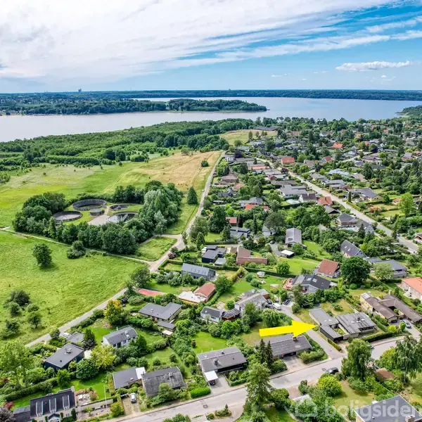 Boligområde med spredte huse markeret med en gul pil, omgivet af grønt landskab. Veje snor sig gennem bebyggelsen med stor sø i horisonten under blå himmel med skyer.