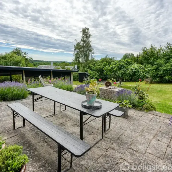 En sort træbænk og bord står på en flisebelagt terrasse, omgivet af blomster og buske. I baggrunden ses en grønt område og træer under en overskyet himmel.